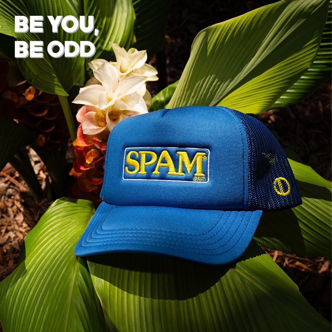 A blue cap with "SPAM" in yellow letters is set among tropical leaves and flowers. The text "BE YOU, BE ODD" appears in bold white letters in the corner.