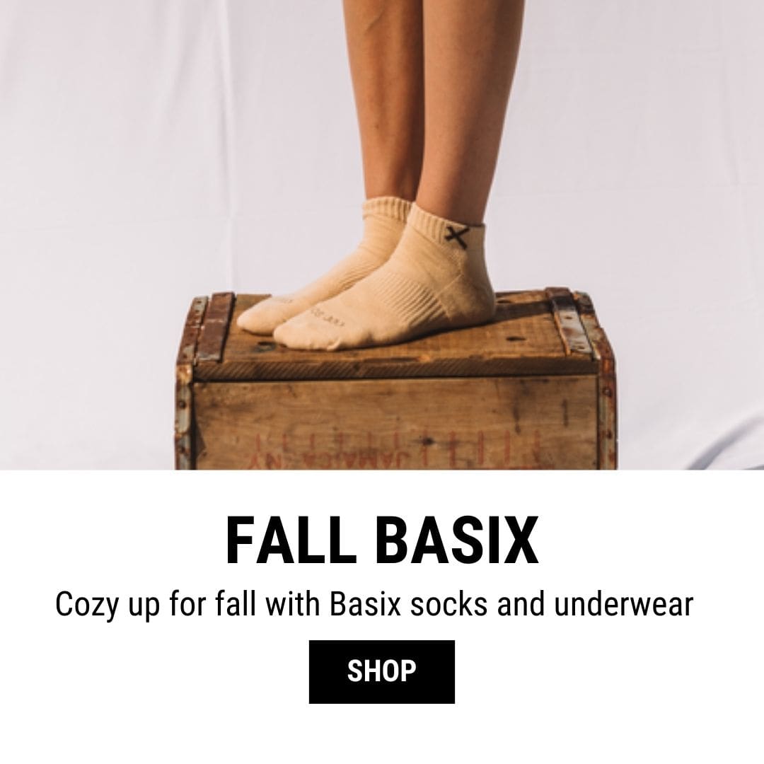 A pair of feet wearing beige socks with a simple "X" logo near the ankle, standing on a vintage wooden box. The background is a plain white surface. The text below reads "FALL BASIX" and "Cozy up for fall with Basix socks and underwear." A "SHOP" button is displayed beneath the text.