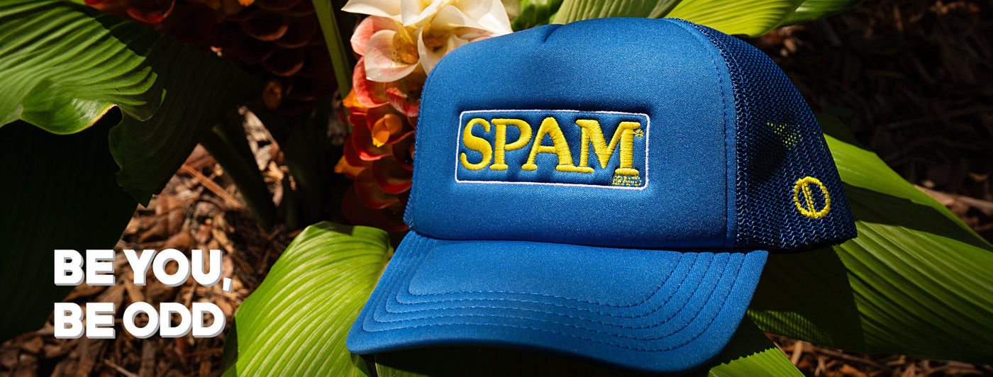 A blue cap with "SPAM" in yellow letters is set among tropical leaves and flowers. The text "BE YOU, BE ODD" appears in bold white letters in the corner.